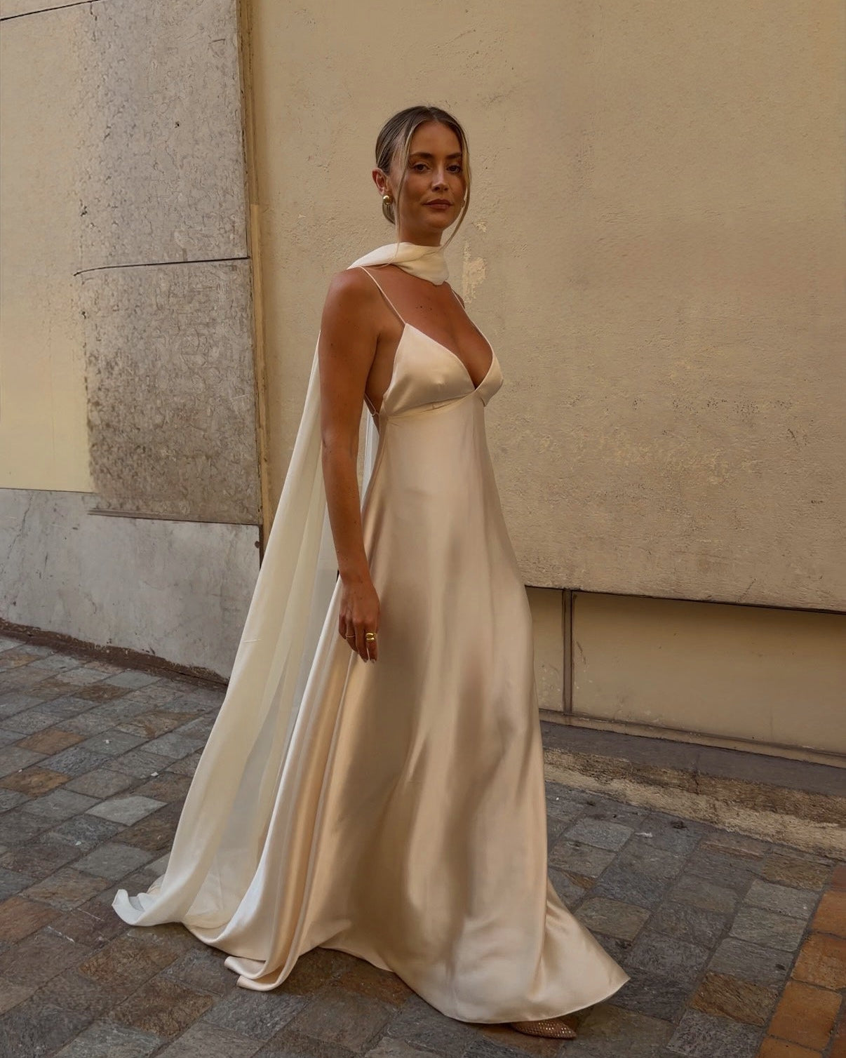 A woman wearing a flowing champagne satin wedding gown with a draped cape-style overlay, standing on a cobblestone street against a beige wall.
