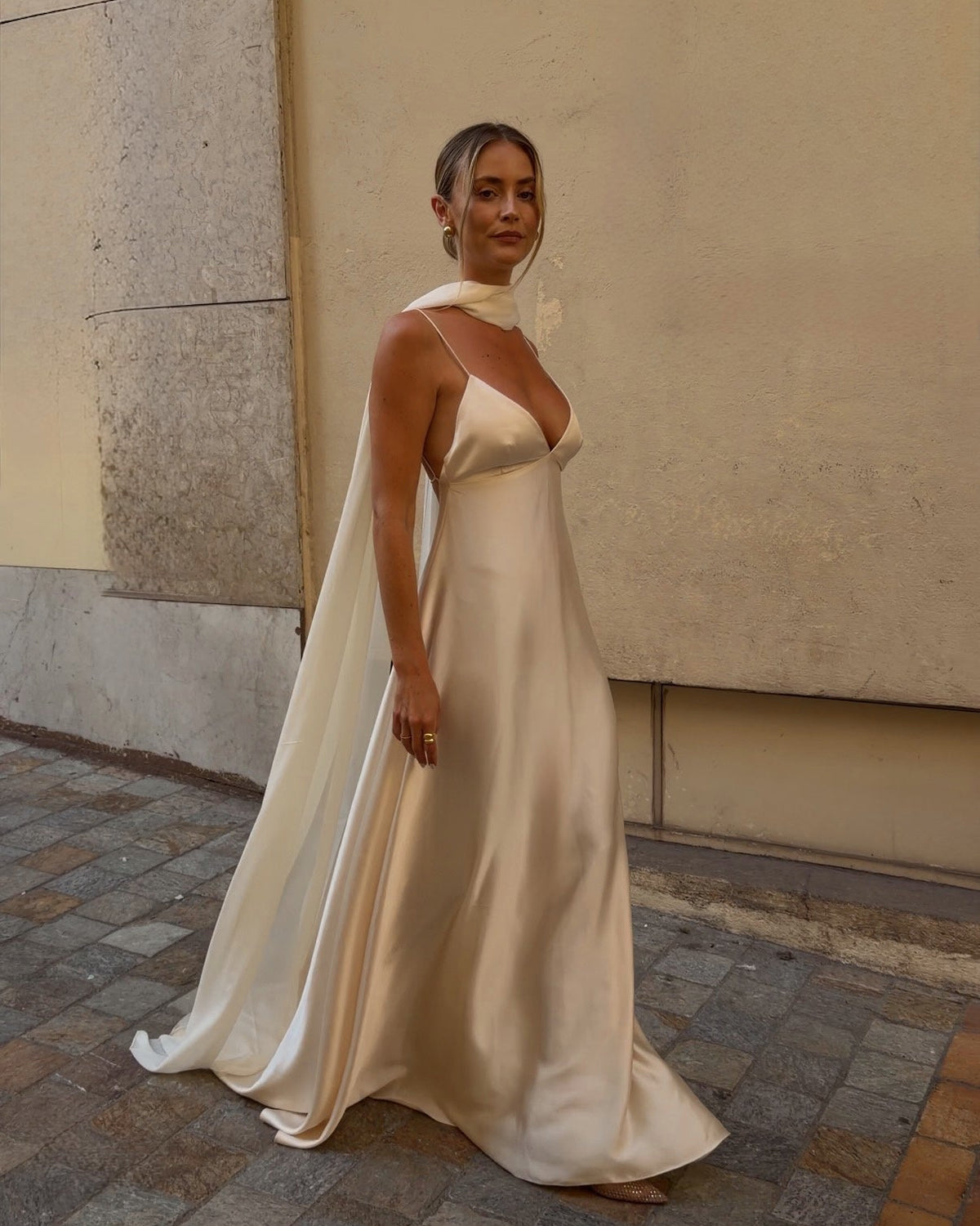 A woman wearing a flowing champagne satin wedding gown with a draped cape-style overlay, standing on a cobblestone street against a beige wall.