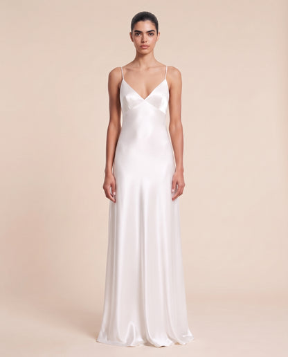 A woman modeling a floor-length white satin slip dress with thin spaghetti straps and a V-neckline, photographed against a solid warm beige studio background.