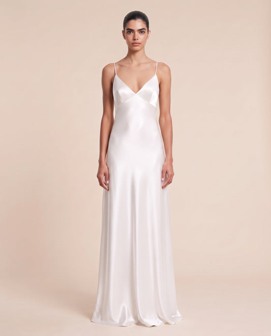 A woman modeling a floor-length white satin slip dress with thin spaghetti straps and a V-neckline, photographed against a solid warm beige studio background.